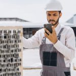 Construction operative in hard hat using smartphone to complete equipment fault reporting on a construction site