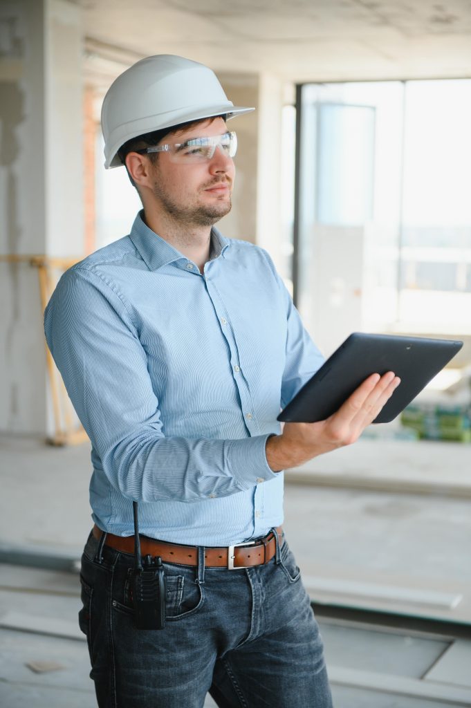 Construction site manager in hard hat and safety glasses reviewing equipment fault reporting data on a tablet inside a building under construction