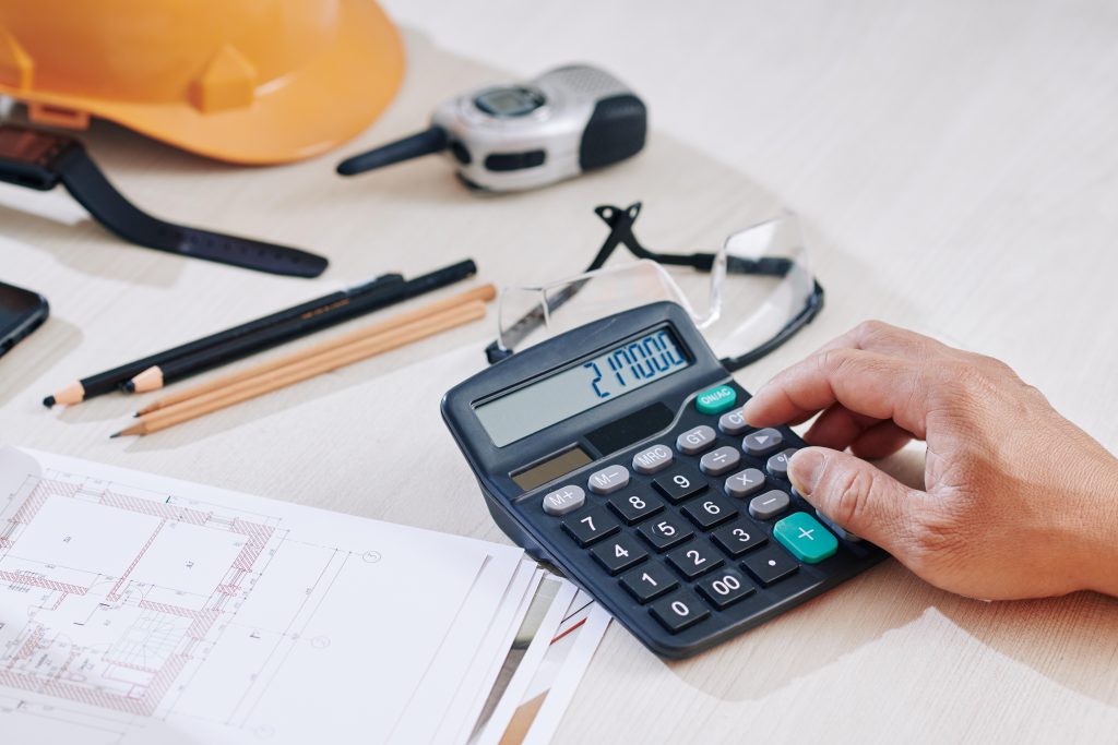 Construction manager calculating fleet inspection software ROI on a desk calculator with hard hat, safety glasses and site blueprints