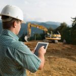Construction site manager reviewing pre-use inspection software on a tablet with an excavator working in the background